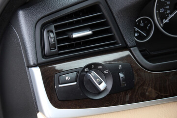 Fototapeta premium Close-up of luxury car dashboard with air vent, headlight control knob, wood trim, and black leather paneling, highlighting premium interior. Premium car light control. Headlight switch, fog lights.