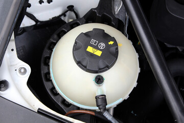 Close-up of modern car coolant reservoir with black cap marked by warning labels and yellow arrows, showing essential engine maintenance component under hood. Car cooling system.