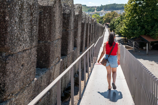 Caminhada solarenga sobre o Adarve da Muralha medieval de Guimar&atilde;es, mostrando uma turista a percorrer o passadi&ccedil;o moderno junto ao antigo muro de pedra, com vegeta&ccedil;&atilde;o e paisagem urbana ao fundo