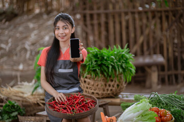 A young woman proudly shows fresh chili peppers on her smartphone in a vibrant market, highlighting agriculture