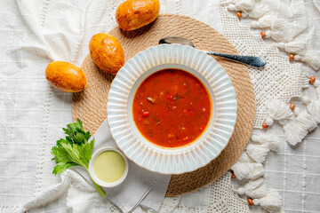 A bowl of rich tomato soup sits on a woven placemat, accompanied by golden bread and fresh herbs