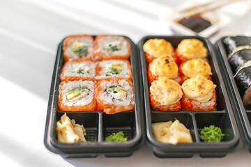 Fresh sushi rolls assortment in black takeout box with salmon avocado cream cheese baked rolls with sauce served with wasabi and ginger on white background
