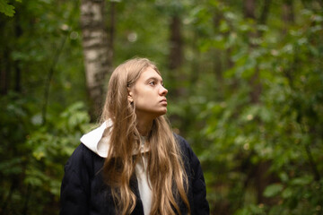 Naklejka premium A young woman with long hair stands in a green forest, looking up thoughtfully. Tall trees and greenery surround her, creating a peaceful atmosphere in the afternoon light.