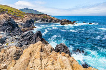 Dramatic Big Sur coastline along Highway 1, California. Waves crash against rugged cliffs, forming turquoise patterns in the Pacific Ocean, showcasing the raw beauty of the California coast