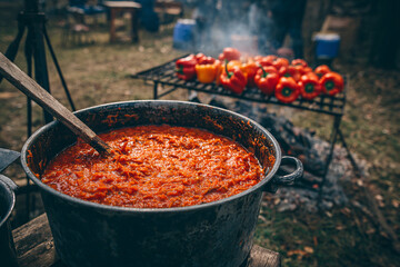 Large pot of simmering ajvar with grilled peppers in the background, outdoor cooking.