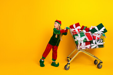 Fototapeta premium Adorable child dressed as an elf pushes a cart full of holiday gifts on a bold yellow background, symbolizing Xmas magic
