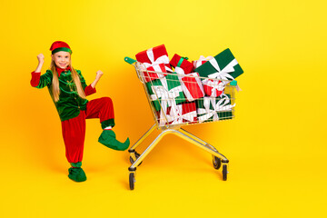 Festive young child dressed as a holiday elf poses joyfully with a cart full of wrapped gifts against a yellow background
