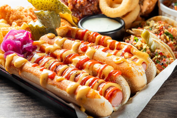 Assorted fast food on tray including hot dogs with mustard and ketchup, tacos, onion rings, pickles, and dipping sauce. Vibrant colors and appetizing street food presentation.