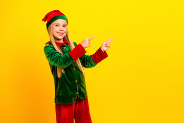 Smiling young girl dressed as a Christmas elf against a vibrant yellow background, cheerfully pointing with festive enthusiasm.
