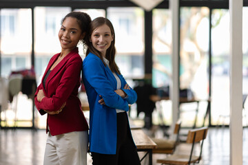 Businesswoman smiling with arms crossed in office. Two confident businesswomen in a modern office, arms crossed, wearing colorful suit, showing teamwork and success.