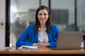Businesswoman smiling with crossed arms in busy office. Businesswoman is smiling while sitting in her company's office.  She is wearing a blue jacket and has her arms crossed.