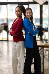 Businesswoman smiling with arms crossed in office. Two confident businesswomen in a modern office, arms crossed, wearing colorful suit, showing teamwork and success.