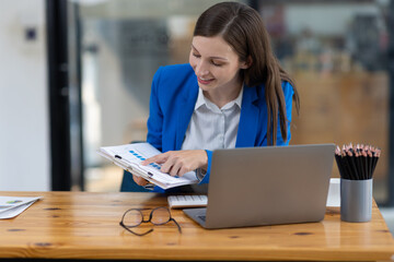 Businesswoman taking notes while having video call on laptop in office. Confident woman smiling during video call in modern office, taking notes. Professionalism and dedication in remote work era.