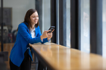Businesswoman using smartphone and looking through office window. Happy woman taking a break in a modern office, using her smartphone and looking out the window.