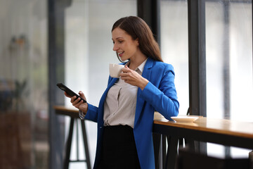 Businesswoman using smartphone and looking through office window. Happy woman taking a break in a modern office, using her smartphone and looking out the window.