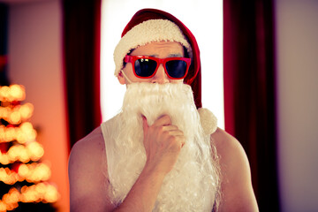 Modern festive Santa Claus with sunglass posing indoors near Christmas tree during the holiday...
