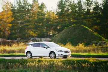 Fototapeta premium Modern white compact hybrid car driving on asphalt road in front of autumn trees