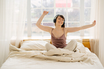 Happy young woman enjoying music with headphones and dancing in bed after waking up. Morning energy, positive emotions, lifestyle and leisure at home.