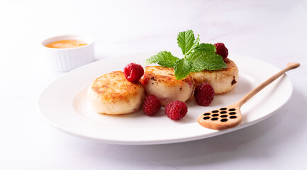 Cottage cheese pancakes with honey, raspberry and mint leaves on white ceramic plate on white marble table close up. Healthy homemade dessert.