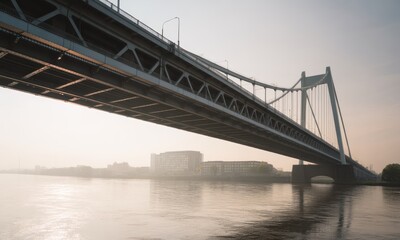 A wide, steel suspension bridge stretches over calm water, with buildings on the far bank in haze