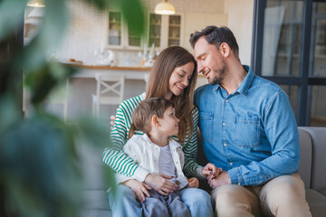 Obraz premium Portrait of happy young Caucasian Family of three with little son relaxing together at home, mom dad and male child looking to each others while embracing on couch in living room