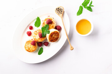 Cottage cheese pancakes with honey, raspberry and mint leaves on white ceramic plate on white marble table top view. Healthy homemade dessert.