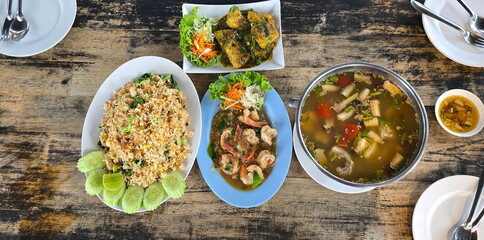 Thai food set served on wooden table with white plates and cutlery. Includes pork fried rice, shrimp stir-fried with shrimp paste, fish tom yam soup, fried fish with chili sauce  fish sauce with chili