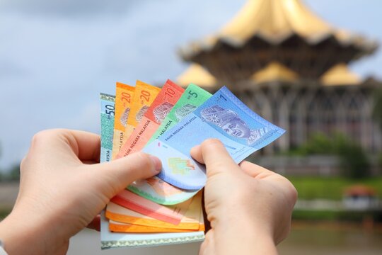 Hand holding Malaysian ringgit paper money with Kuching city skyline in background.