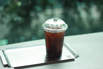 A clear plastic cup filled with iced coffee sits on a gray stone surface. Condensation forms on the outside of the cup, highlighting the cold beverage, with a blurred green natural background behind.