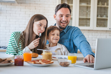Joyful family using laptop during the breakfast in the kitchen watching cartoon have video call, everyone is happy and smiling, parents enjoy spending time with their child kid son little boy