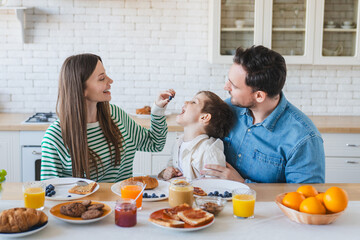 Young happy beautiful family having breakfast together at home. Father, mother and cute little kid son eating healthy food in morning, talking and smiling while sitting in modern kitchen