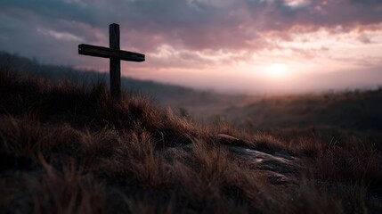 A solitary wooden cross stands on a grassy hilltop bathed in the warm light of sunrise with a misty atmospheric landscape