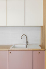 Light wooden table top, white sink with water tap, against a white wall, pink and white kitchen cabinets