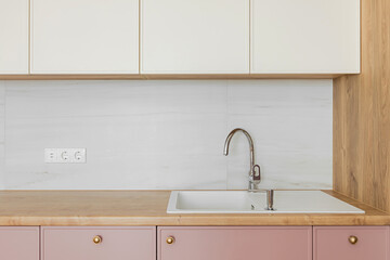 Light wooden table top, white sink with water tap, against a white wall, pink and white kitchen cabinets