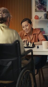Smiling aged woman playing chess with female friend in wheelchair, leisure time