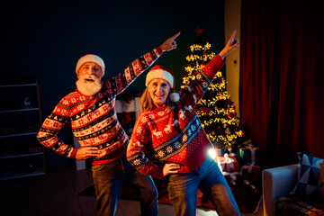 Fototapeta premium Two seniors couple in festive sweaters pose by a Christmas tree at home with gifts and warm lights for a joyful holiday memory