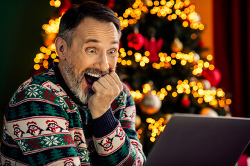 Excited mature man in festive sweater browsing laptop near Christmas tree in cozy home setting