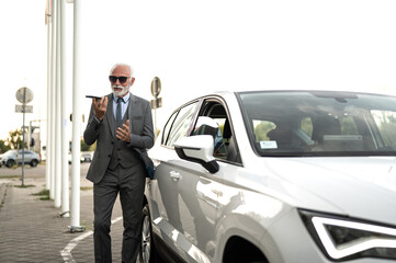 Senior businessman using smartphone voice assistant near car