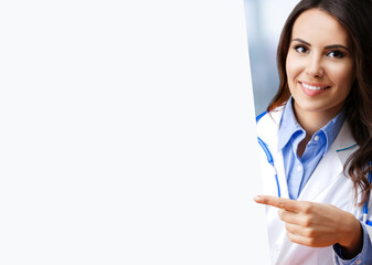 Portrait image of happy smiling female doctor pointing blank signboard with free space for ad slogan text. Young brunette beautiful cute woman in medical, clinic, healthcare concept