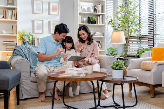 Happy Asian family using tablet,laptop for video call,playing game in the living room and looking at the camera.Smiling parents,their daughter are using a digital tablet for learning or entertainment
