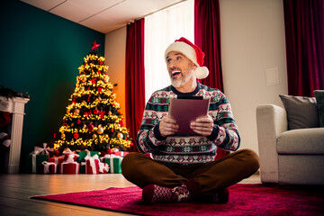 Joyful senior man in festive sweater using tablet while enjoying Christmas atmosphere by the tree