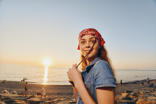 smiling woman wearing red headscarf enjoys sunset on rocky beach holding stick in warm summer evening light - Powered by Adobe