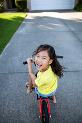 Joyful young asian girl riding a red bicycle on a sunny suburban driveway, laughing and looking back, Generative AI