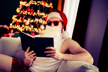 Santa Claus in sunglass reading a book beside a Christmas tree in festive pajamas, enjoying the...