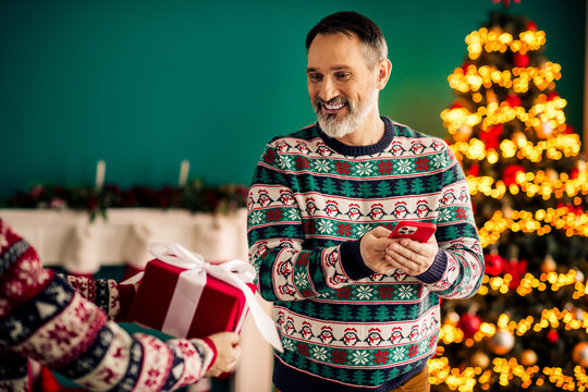 Festive moment with a smiling man holding a smartphone, receiving a gift in a cozy decorated home during Christmas - Powered by Adobe