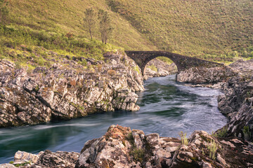 Stone Charcos Bridge over the Isna stream between Sertã and Vila de Rei, Portugal, surrounded by rugged rocks and hills in a stunning natural setting during the soft light of late afternoon.