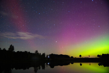 Brilliant purple and green aurora glows above a starry night sky, mirrored on a calm lake with...