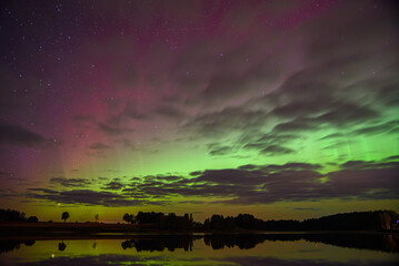 Green and purple aurora lights glow through a starry night sky with scattered clouds, reflecting beautifully on a still lake below.