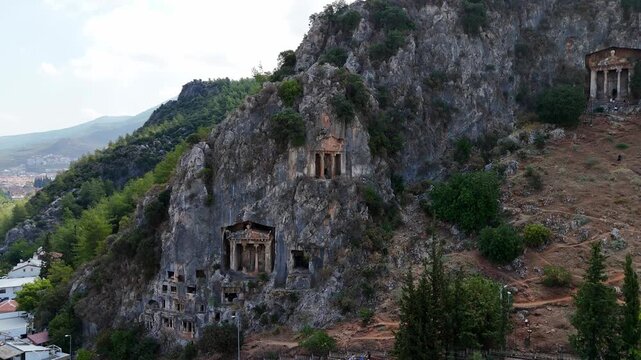 Aerial view of ancient Lycian rock tombs carved into the cliffs of Telmessos in Fethiye, Turkey, surrounded by mountains and greenery, showcasing historical and archaeological heritage.