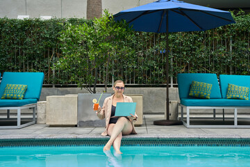 Businesswoman in sunglasses works on laptop by the poolside while enjoying a refreshing cocktail. Concept of digital nomad lifestyle, summer productivity, remote work, and luxury outdoor leisure.